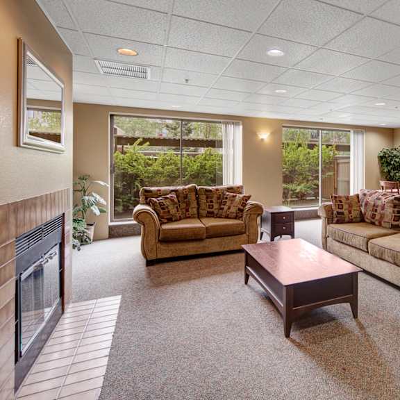 spacious resident lounge here at susitna ridge apartments with two beige sofas, a wooden coffee table, and a fireplace. large windows reveal lush greenery. dining area with a table, chairs, and a potted plant. cozy and inviting tone.