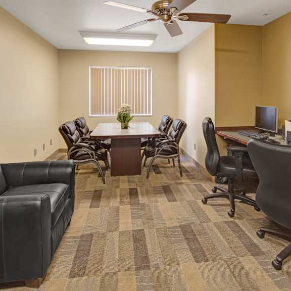 susitna ridge apartment's modern, neutral-toned office features a round table with six leather chairs, a black sofa, two computers on desks, and a window with blinds.