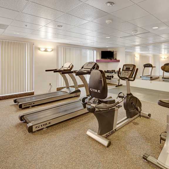 bright gym here at susitna ridge apartments with two treadmills, exercise bike, and elliptical on carpeted floor. mirrors and windows create an open, airy feel. dumbbells rest on a rack.