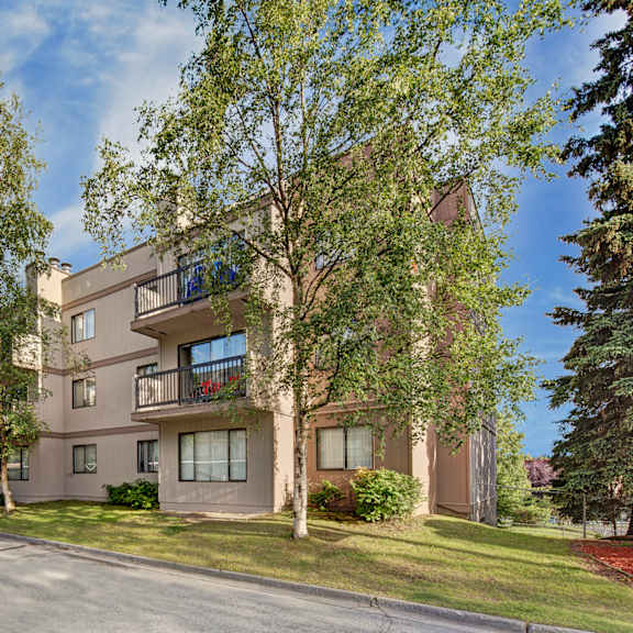 three-story beige sustitna ridge apartment building surrounded by lush green trees under a clear blue sky. balconies feature potted plants, creating a serene atmosphere.