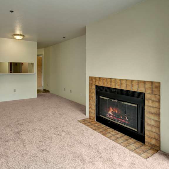 empty living room here at susitna ridge apartments with beige carpet, featuring a lit fireplace with a brown tile surround on the right, and a small serving hatch on the left wall. cozy ambiance.