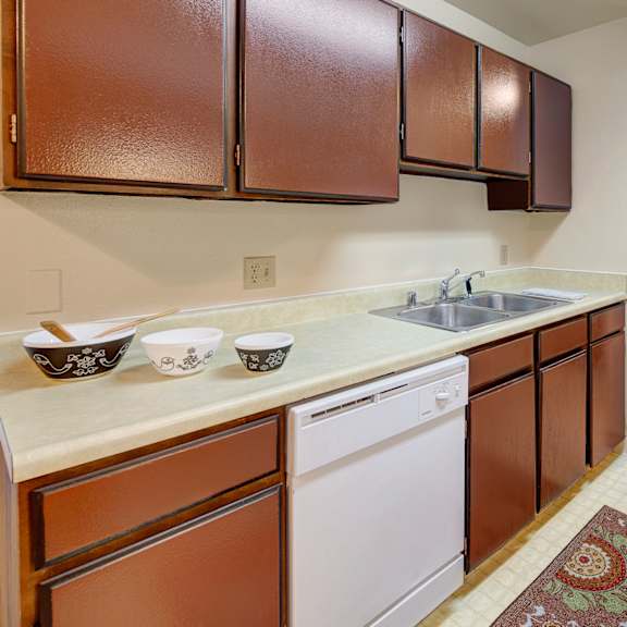 a kitchen here at taiga apartments with dark brown cabinets and a light countertop. three decorative bowls and a wooden spoon sit by the sink. there's a patterned rug on the beige floor.