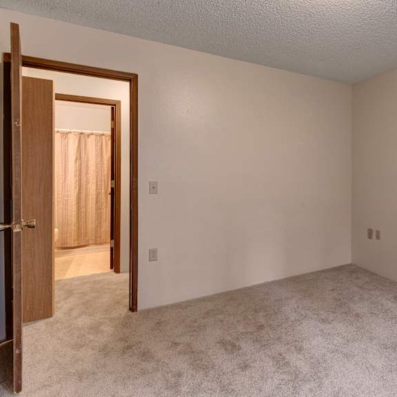 empty beige carpeted bedroom here at taiga apartments with a doorway to a bathroom, partially visible beige shower curtain. neutral tones convey a calm, minimalist atmosphere.