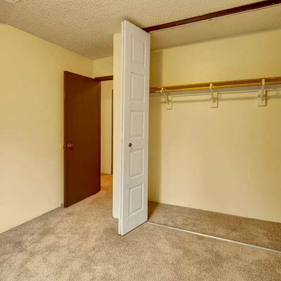 empty beige room here at taiga apartments with open closet doors, revealing a wooden rod and shelf inside. carpeted floor and an open brown door leading to another room. quiet and clean atmosphere.