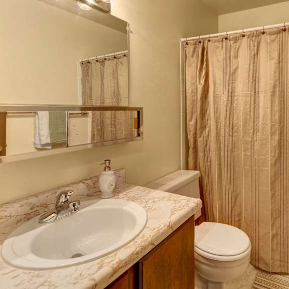 a small bathroom here at taiga apartments with a marble-patterned sink, soap dispenser, and toothbrush holder. beige shower curtain, wooden door with towels, bright lighting.