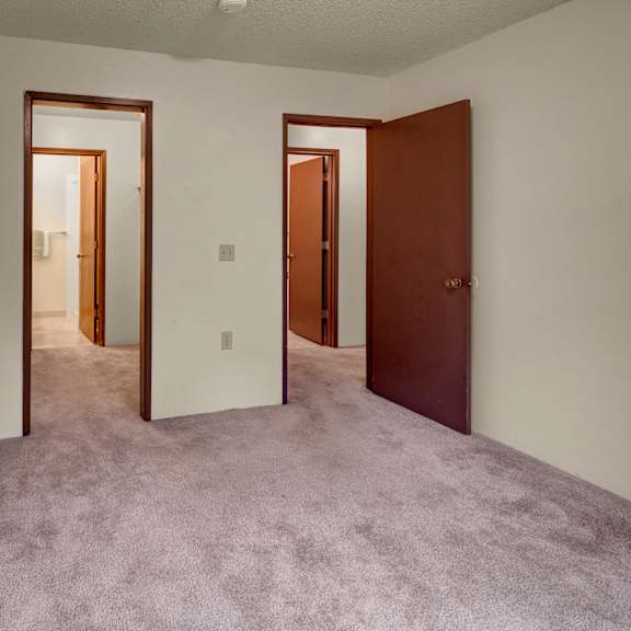 empty bedroom here at taiga apartments with beige carpet, white walls, and open brown doors leading to two additional rooms. the atmosphere is neutral and minimalistic.