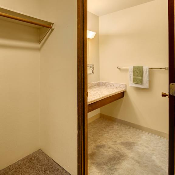 minimalist interior here at taiga apartments showing an empty closet with a wooden rod and shelf. adjacent bathroom features a marble countertop and towel rack with towels.