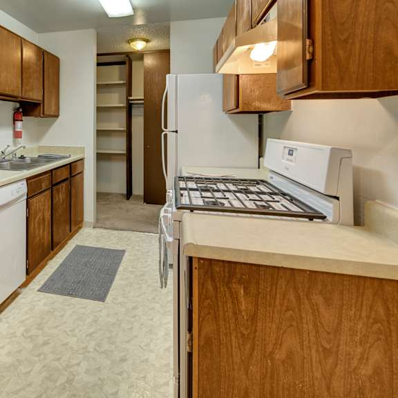 narrow kitchen here at taiga apartments with wooden cabinets, beige countertops, a gas stove, white appliances, and a sink. a pantry, grey rug, and scattered bowls create a cozy feel.