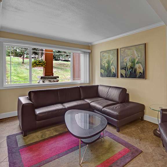 cozy living room here at taiga apartments with brown leather sectional and armchair, a glass coffee table on a colorful rug, large window with garden view, and floral paintings on yellow walls.