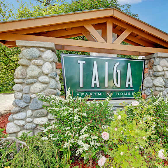 stone sign for taiga apartment homes amid lush green foliage and blooming flowers, set against a bright blue sky, conveying a welcoming and serene atmosphere.