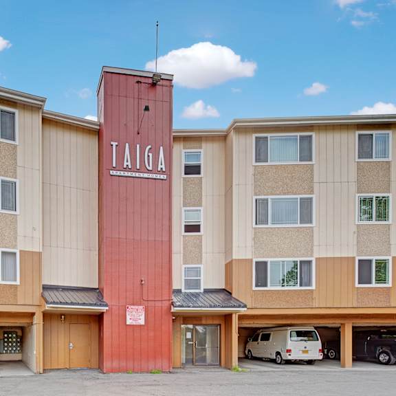 four-story taiga apartment building with a vertical red sign reading "taiga." beige facade with multiple windows and cars parked in ground floor garages.