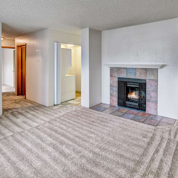 a spacious living room here at terrace on the lake apartments with beige carpet features a lit fireplace with a tiled mantel. the room is bright, with white walls and a cozy feel.