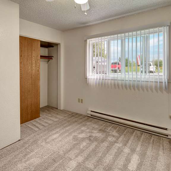 empty bedroom terrace on the lake apartments with beige carpet, white walls, and vertical blinds on a window. an open closet with a wooden door is on the left, creating a neutral, clean atmosphere.