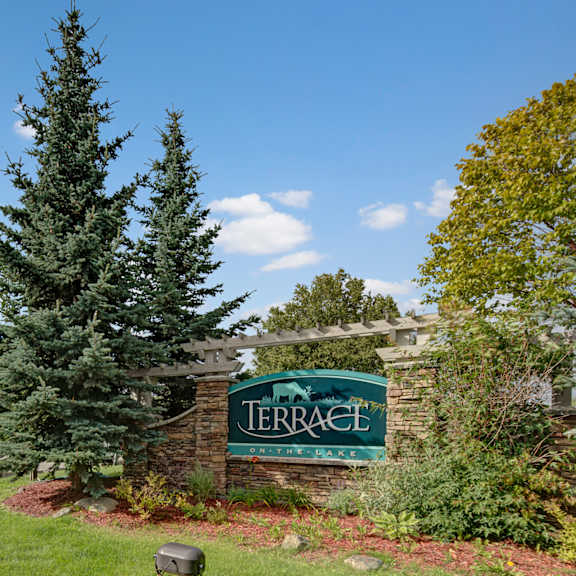 apartment complex entrance with stone sign reading "terrace on the lake," flanked by tall evergreen trees. a building and parked cars are visible under a bright blue sky.