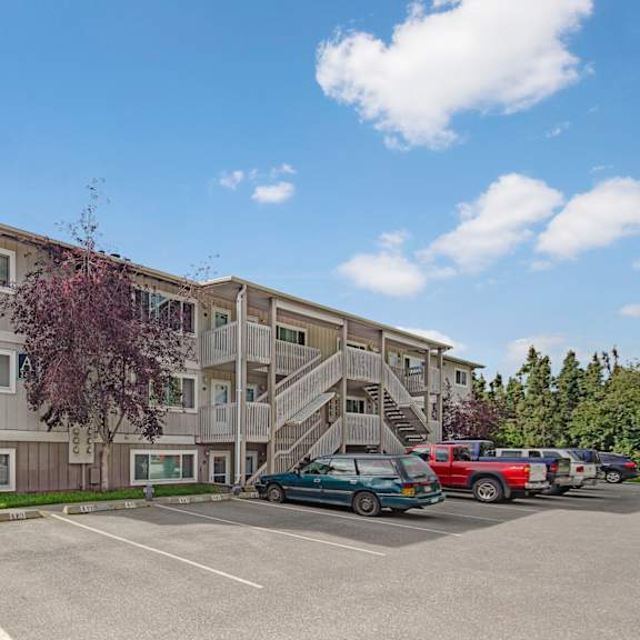 three-story beige terrace on the lake apartment building with external staircases, parked cars in front, and a cloudy blue sky. trees frame the scene, offering a serene feel.