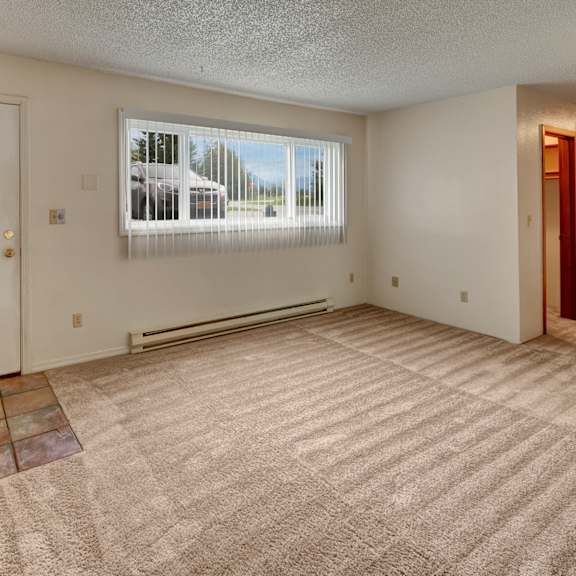 empty living room here at terrace on the lake apartments with beige carpet, white walls, and a light blue door with windows at the top. large window with vertical blinds lets in natural light.