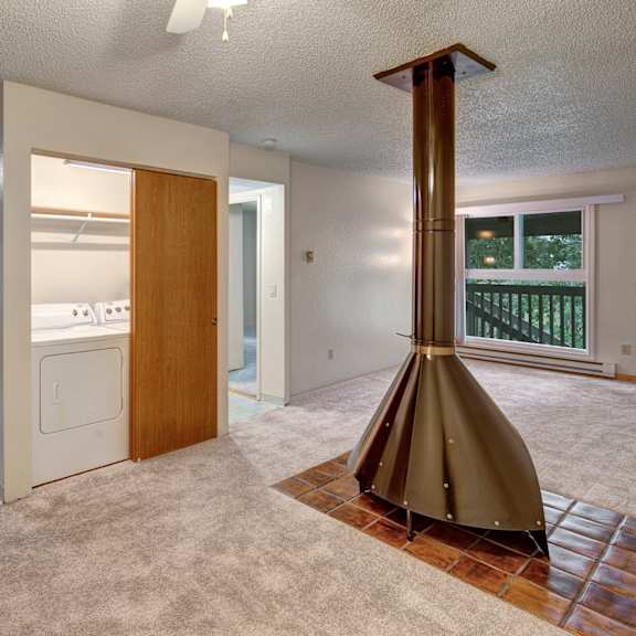 open-plan living space here at terrace on the lake apartments with beige carpet, a central vintage cone fireplace, and a windowed door leading to a balcony. adjacent is a utility room with a washer and dryer.