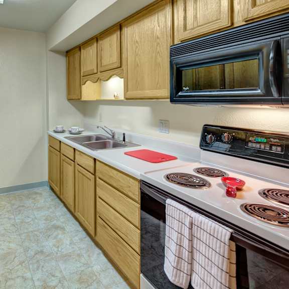 a narrow kitchen here at terrace on the lake apartments with wooden cabinets, a double sink, stove, and microwave. a red cutting board and striped towels add a cozy touch. bright lighting.