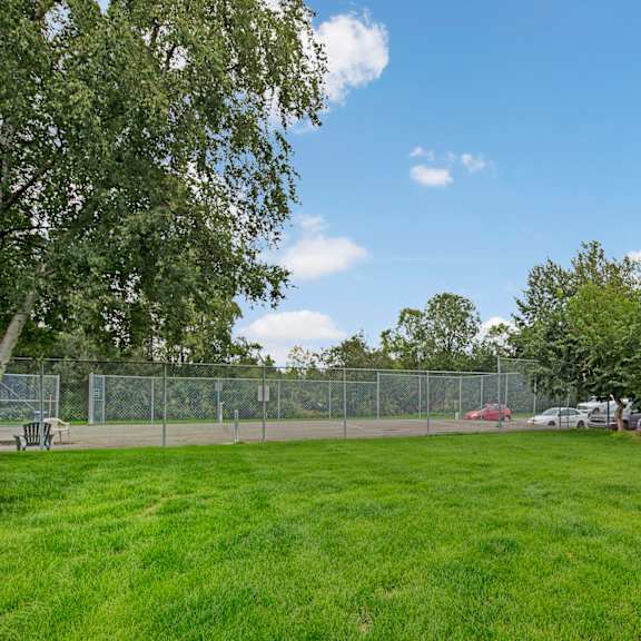lush green lawn in the foreground, fenced tennis court to the left, and parked cars to the right. clear blue sky and leafy trees create a serene atmosphere here at terrace on the lake apartments.