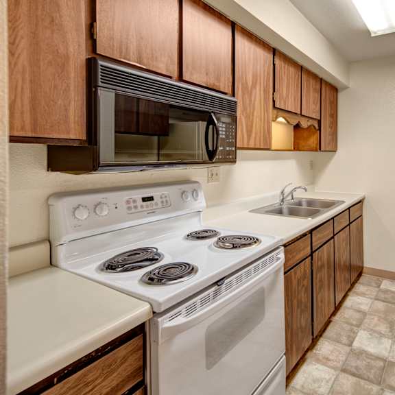 narrow kitchen here at terrace on the lake apartments with brown wood cabinets, white appliances, and stone-patterned linoleum floor. features a stove, microwave, sink, and fridge.