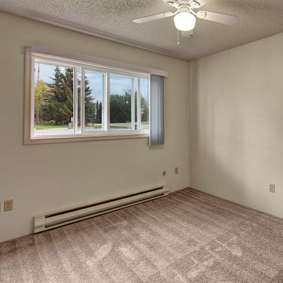empty bedroom here at terrace on the lake apartments with beige carpet and pale walls, featuring a large window with venetian blinds. a ceiling fan hangs above, and sunlight creates gentle shadows.