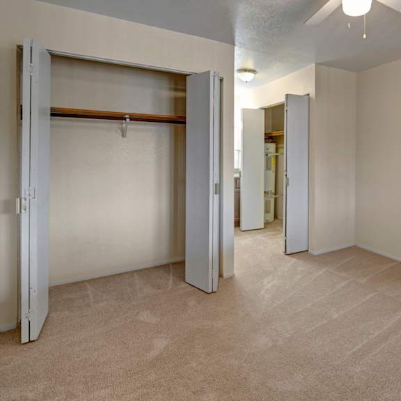 empty beige bedroom here at the club at eagle pointe apartments with open closets revealing a wooden rod and shelves. carpeted floor, a ceiling fan, and soft lighting create a neutral, spacious feel.