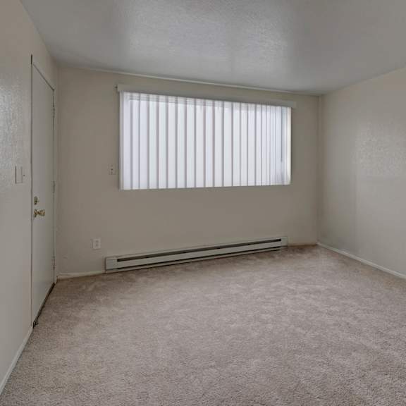 empty, beige-carpeted living room here at the club at eagle pointe apartments with pale walls and a large window with vertical blinds, creating a clean and neutral atmosphere.