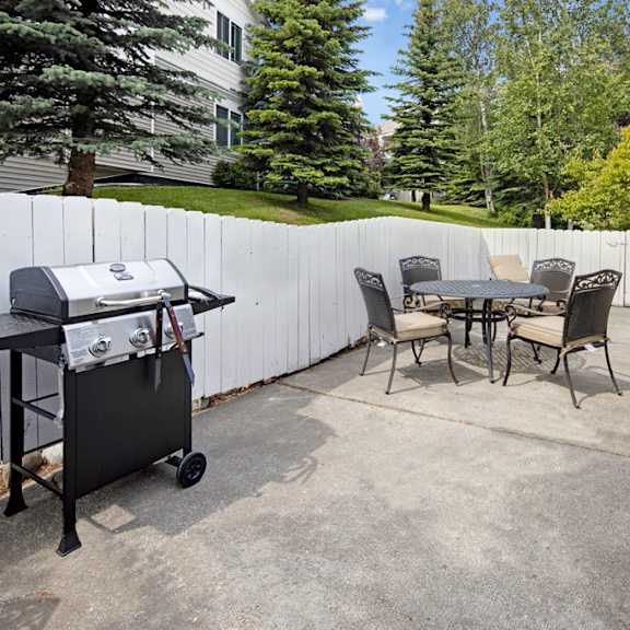 a patio here at the highlands luxury apartments with a barbecue grill and a round table surrounded by four chairs. a white fence and trees surround the area, creating a peaceful setting.