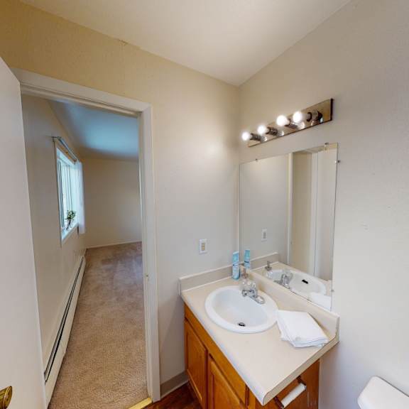 bright bathroom here at the village at calais apartments with beige walls, a wood cabinet sink, and a large mirror. a corridor with carpet and a window leads to another room.