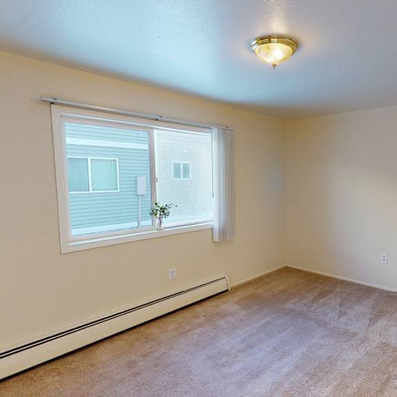 spacious, empty bedroom here at the village at calais apartments with beige carpet and walls, large window with vertical blinds, and a small plant on the sill. overhead light fixture provides a cozy feel.