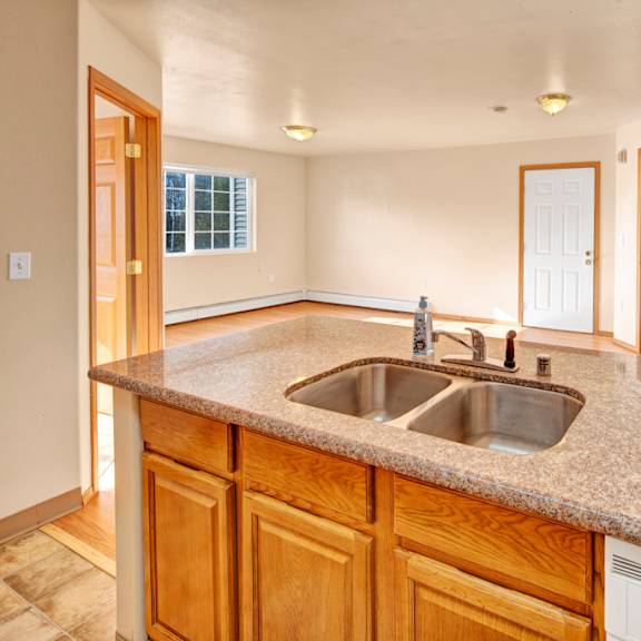 spacious kitchen here at timber ridge apartments with granite countertop and stainless steel sink, overlooking a bright living area. warm lighting and wooden cabinets create a cozy atmosphere.