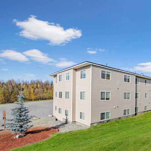 four-story beige timber ridge apartment building under a bright blue sky. surrounding green lawn and trees create a peaceful, suburban setting.