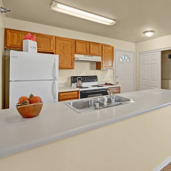 kitchen here at timber ridge apartments with wooden cabinets, white appliances, and a countertop with a sink. a bowl of squash is on the counter. closet with hangers is open.