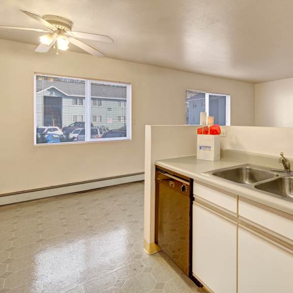 a clean, bright kitchen here at timber ridge apartments with a dual sink, dishwasher, and light countertops. two windows reveal a suburban view. a ceiling fan adds a cozy touch.