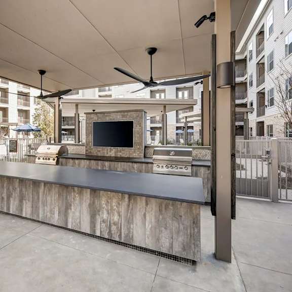 a covered outdoor kitchen area here at alden at cedar park with a long gray counter, built-in stainless grills, mounted tv, ceiling fans, and modern apartment buildings visible around the patio space.
