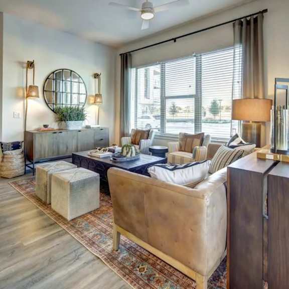 a living room here at alden at cedar park with a brown leather sofa, striped accent chairs, patterned rug, black coffee table, gray ottomans, large bright windows, and open view into a staged bedroom.