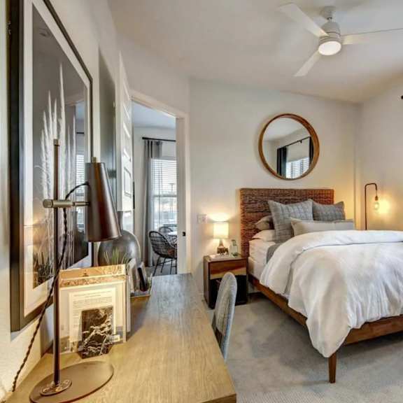 a bedroom here at alden at cedar park with a wood bed frame, woven headboard, white bedding, round wall mirror, warm bedside lamps, desk workspace, and large window with dark blue curtains.