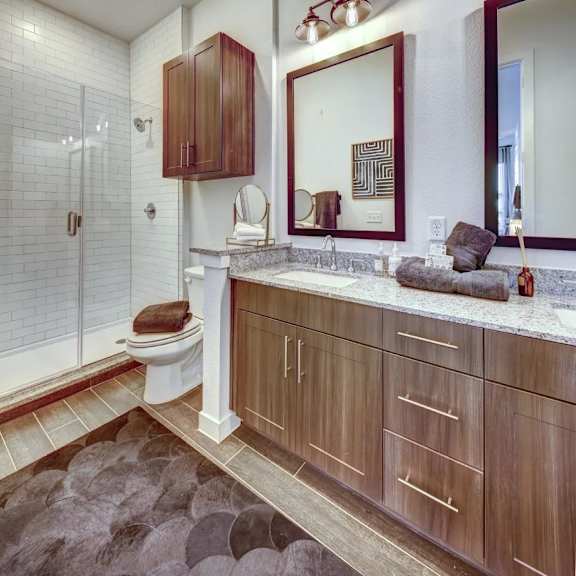 a bathroom here at alden at cedar park with wood-tone vanity cabinets, granite countertop, double mirrors, glass shower with white tile, and dark brown bath rug and towels.