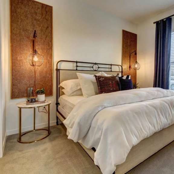 a bedroom here at alden at cedar park with a black metal bed frame, white bedding, dark accent pillows, blue curtains, round gold side table, and warm wall panels with hanging lights.