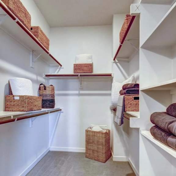 a walk-in closet here at alden at cedar park with white shelves, hanging rods, woven storage baskets, and neatly folded brown towels on open side shelving.