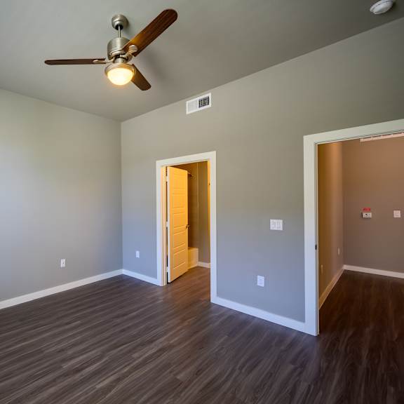 a bright, empty bedroom here at harmony luxury with warm wood-style flooring, soft gray walls, and a ceiling fan with built-in lighting, complemented by natural light from a window and an open doorway leading to an adjoining space for a clean, comfortable feel.