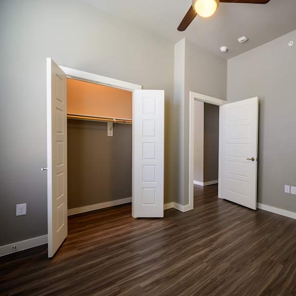a clean, empty bedroom here at harmony luxury with warm wood-style flooring, soft gray walls, and a ceiling fan with built-in lighting, featuring an open walk-in closet with shelving and an open doorway that adds a bright, functional, and comfortable feel.
