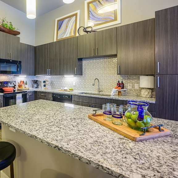 a modern kitchen here at harmony luxury with dark wood cabinetry, granite countertops, and a large center island, featuring sleek black appliances, pendant lighting, a patterned tile backsplash, and bar-style seating that create a polished, upscale atmosphere.