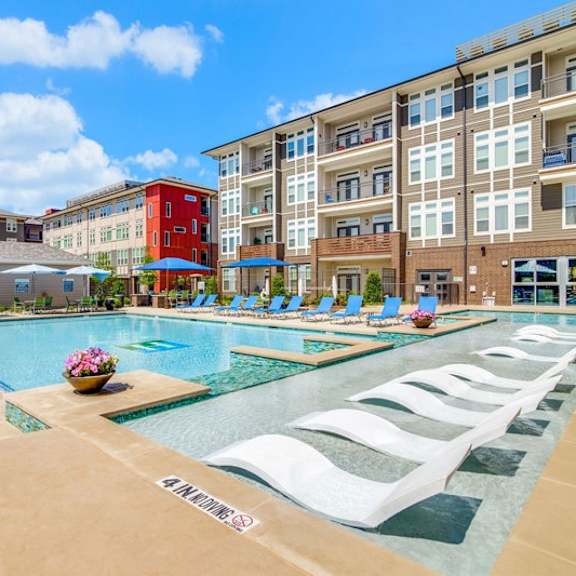 a resort-style outdoor pool here at harmony luxury with in-water lounge chairs, surrounding poolside seating, and modern apartment buildings in the background, offering a bright, relaxing atmosphere designed for leisure and warm-weather enjoyment.