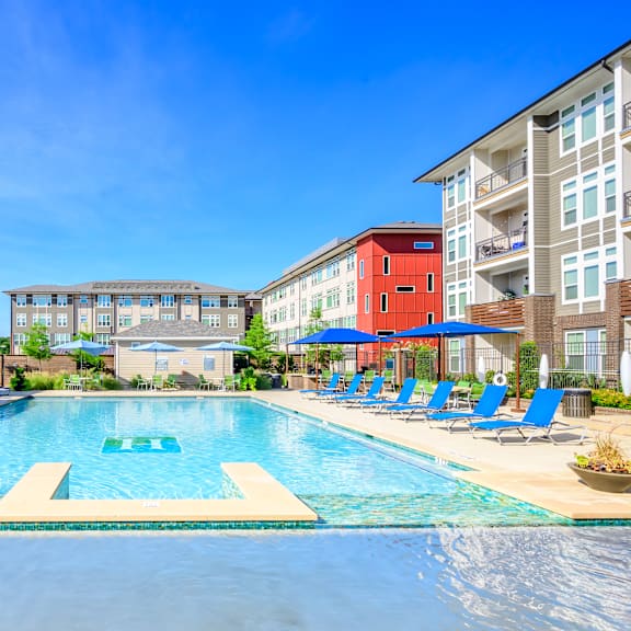 a resort-style swimming pool here at harmony luxury with clear blue water, in-water ledges, poolside lounge chairs, shaded seating areas, and modern apartment buildings surrounding the space, creating a bright and relaxing outdoor retreat.