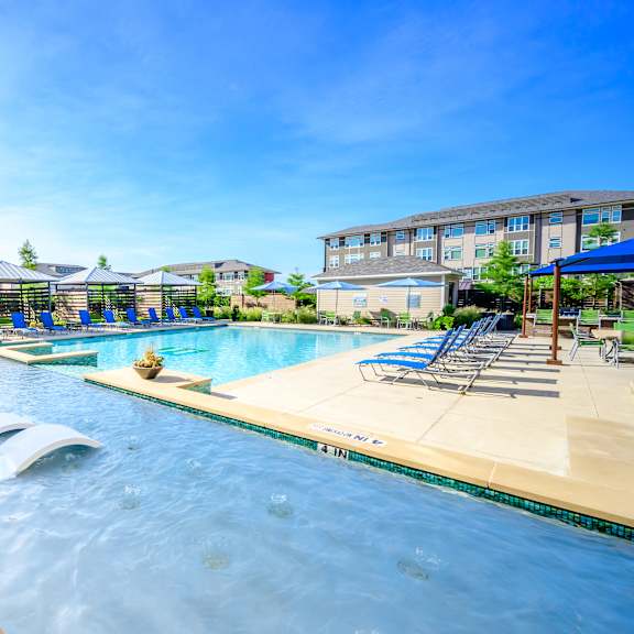 a resort-style swimming pool here at harmony luxury with in-water lounge chairs, clear blue water, poolside seating, and shaded cabanas, surrounded by landscaped greenery and modern apartment buildings for a relaxing, upscale outdoor experience.