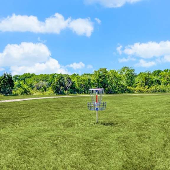 a spacious outdoor green space here at harmony luxury with a well-maintained grassy field, a disc golf basket, and surrounding trees, offering a peaceful, open area for recreation and outdoor activities under a bright blue sky.