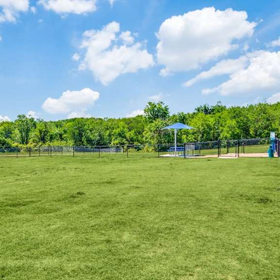 a large outdoor dog park here at harmony luxury with a fenced grassy area, open space for play, shaded seating, and surrounding trees, offering a comfortable and pet-friendly environment for exercise and relaxation.