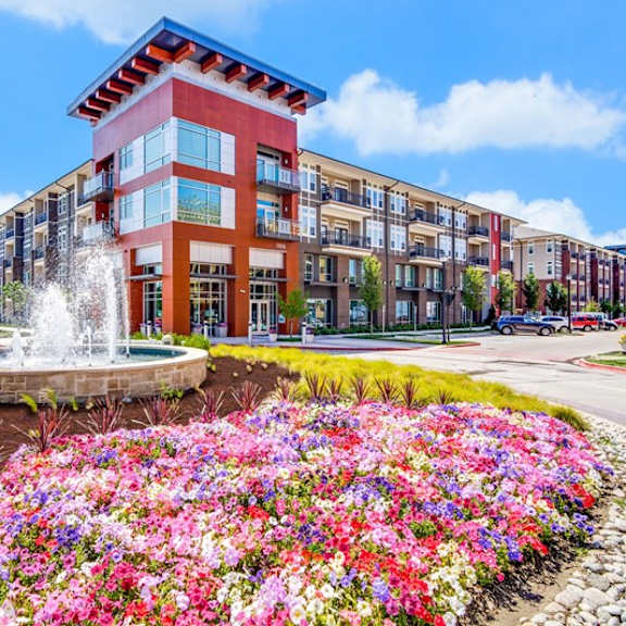 a landscaped exterior view here at harmony luxury featuring a decorative fountain, vibrant flower beds, and modern apartment buildings with balconies, creating a colorful and inviting entrance to the community.