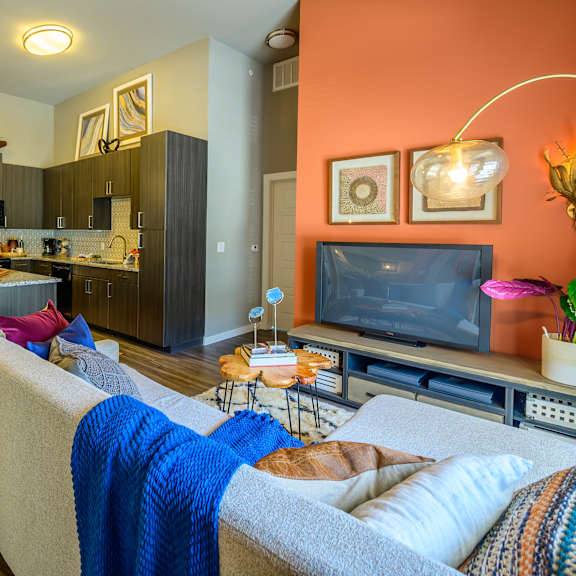 a vibrant open-concept living space here at harmony luxury with a cozy sectional sofa, colorful accent pillows, and a media console, flowing into a modern kitchen with dark cabinetry, granite countertops, pendant lighting, and bar seating that create a stylish, upscale atmosphere.